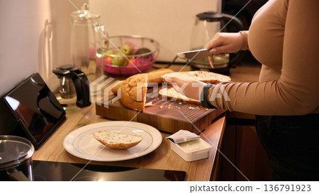 Woman cutting loaf and placing slice on plate near tablet on kitchen counter. Woman cutting loaf and placing slice on plate near tablet on kitchen counter. 136791923