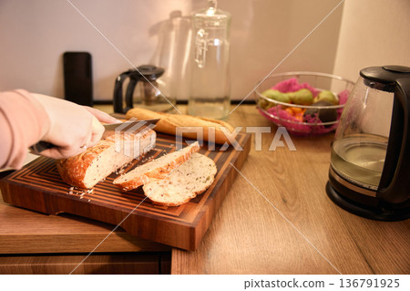 Close up of hands cutting fresh bread on wooden board in contemporary kitchen Close up of hands cutting fresh bread on wooden board in contemporary kitchen 136791925