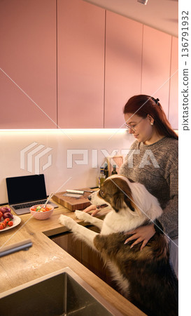 Woman standing with dog near countertop, laptop and fresh salad in modern kitchen. Woman standing with dog near countertop, laptop and fresh salad in modern kitchen. 136791932