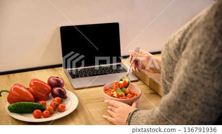 Woman holding bowl of fresh salad with laptop on countertop in contemporary kitchen. Woman holding bowl of fresh salad with laptop on countertop in contemporary kitchen. 136791936