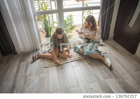 A mother and her daughter sit on a rug with hamster in a well-lit room. They are focused on a phone A mother and her daughter sit on a rug with hamster in a well-lit room. They are focused on a phone 136792357