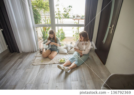 girl and women relax on a rug in a modern indoor space. They share snacks while sitting  136792369