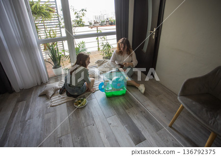 Two girls share snacks and play with a pet on a rug inside a bright room with large windows  136792375