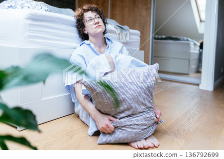 Middle aged white woman with curly hair and glasses sitting barefoot on bedroom floor, hugging pillow, looking forward with indifferent facial expression, emotional numbness, depression concept Middle aged white woman with curly hair and glasses sitting barefoot on bedroom floor, hugging pillow, looking forward with indifferent facial expression, emotional numbness, depression concept 136792699