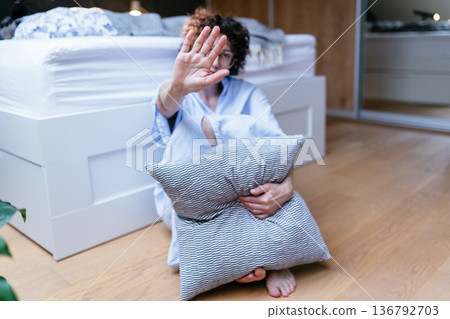 Middle aged curly haired woman sitting barefoot on floor, holding pillow tightly, raising hand in stop gesture, serious facial expression, emotional distress, depression concept, private home interior Middle aged curly haired woman sitting barefoot on floor, holding pillow tightly, raising hand in stop gesture, serious facial expression, emotional distress, depression concept, private home interior 136792703