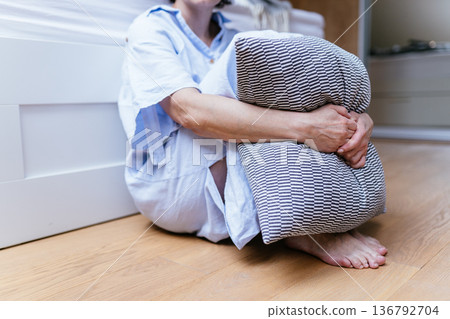 Close view of unrecognizable middle aged woman sitting on bedroom floor barefoot, tightly hugging pillow, emotional distress, depression, anxiety, loneliness concept, private home interior. Close view of unrecognizable middle aged woman sitting on bedroom floor barefoot, tightly hugging pillow, emotional distress, depression, anxiety, loneliness concept, private home interior. 136792704