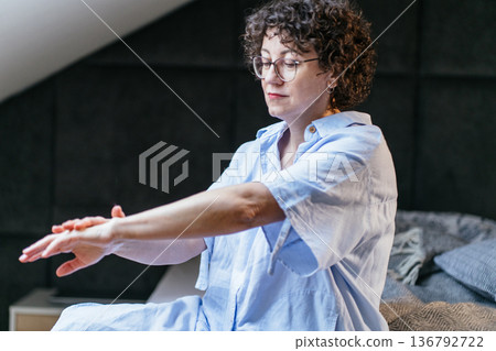 Thoughtful mature middle aged White female wearing glasses sits barefoot on bed hugging pillow. Reflective mood, emotional pause after work, mental health concept, quiet evening at home. Thoughtful mature middle aged White female wearing glasses sits barefoot on bed hugging pillow. Reflective mood, emotional pause after work, mental health concept, quiet evening at home. 136792722