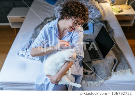Warmhearted White female in her 40s wearing glasses, smiling while petting white cat on bed near open laptop. Cozy bedroom, soft blanket, home office break, pet friendly work, calm weekend morning . Warmhearted White female in her 40s wearing glasses, smiling while petting white cat on bed near open laptop. Cozy bedroom, soft blanket, home office break, pet friendly work, calm weekend morning . 136792732