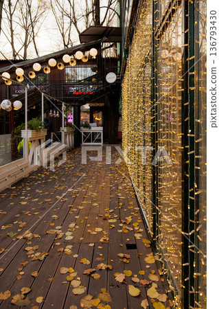 Golden leaves dance along a cozy walkway adorned with twinkling lights in autumn's gentle embrace 136793430