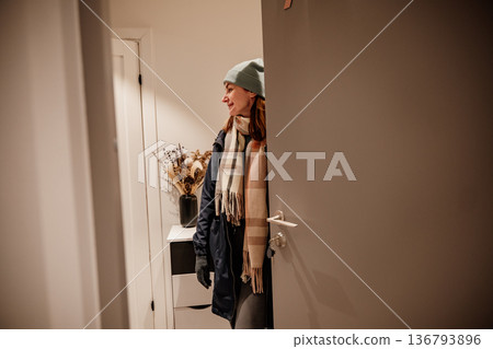 Woman opens door and smiles while wearing warm clothes in a cozy home setting Woman opens door and smiles while wearing warm clothes in a cozy home setting 136793896