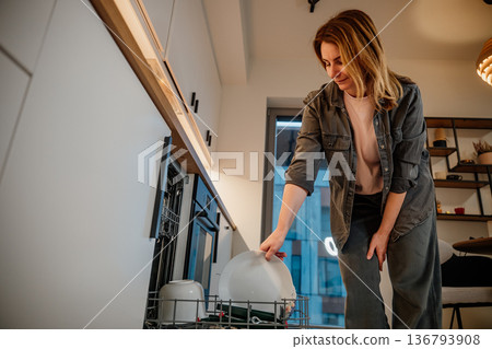 Woman loading clean dishes into modern kitchen dishwasher during the day 136793908