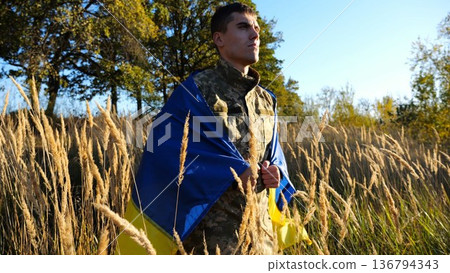 Military man in uniform stands with flag of Ukraine on shoulders at countryside. Portrait of male ukrainian soldier with national banner in honor of victory against russian aggression. End of war Military man in uniform stands with flag of Ukraine on shoulders at countryside. Portrait of male ukrainian soldier with national banner in honor of victory against russian aggression. End of war 136794343