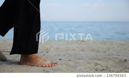 Legs of adult woman stepping along sea shore. Female feet walking on sand at the beach with seascape at background. Barefoot lady strolling near ocean coast. Concept of summer vacation. 136794363