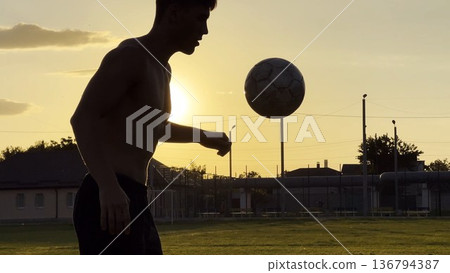 Young man juggling soccer ball on stadium at sunset. Professional footballer kicking ball at green field. Sportsman practicing tricks at meadow with sunlight at background. Freestyle football 136794387