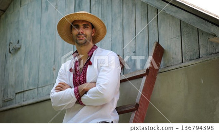 Portrait of smiling guy in straw hat against the background of his house. Young ukrainian man wearing an embroidered shirt looking into camera outdoor. Concept of national identity and patriotism 136794398