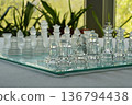 Glass chess pieces on a glass board in a room against the window with green plants and summer view. Close up. Copy space at the bottom. The concept of hobby and leisure. Real photo shot at eye level. 136794438