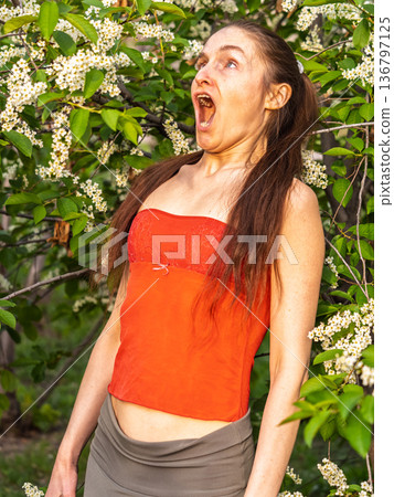 Young woman sneezes in the park against the background of a flowering tree. Allergy to pollen concept. Young woman sneezes in the park against the background of a flowering tree. Allergy to pollen concept. 136797125