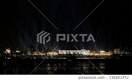 golden lights illuminate waterfront scene, spectacular illumination enhances lively urban riverside spectacle, dazzling beam arrangement transforms city skyline and riverfront into vibrant celebration 136801452