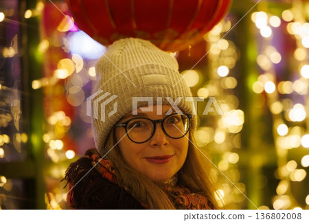 Smiling woman wearing warm hat and scarf beneath colorful lights during festive celebration in the evening 136802008