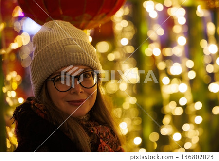 Smiling woman in cozy hat stands among colorful lights and lanterns during festive celebration at night Smiling woman in cozy hat stands among colorful lights and lanterns during festive celebration at night 136802023