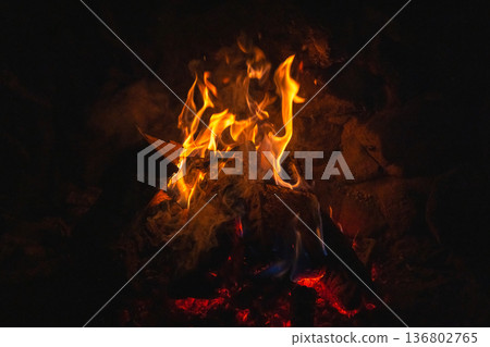 A glowing campfire burns in the dark, casting amber light and blue flames. Charred logs crackle as embers glow, creating a warm, intimate outdoor scene ideal for camping and rustic gatherings 136802765