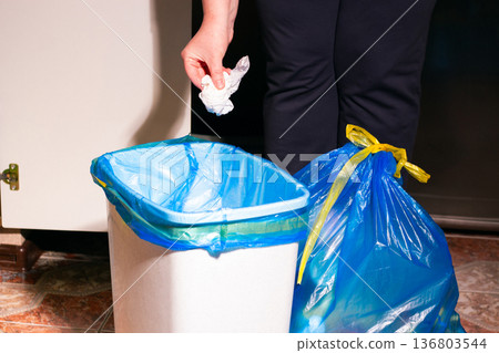Hand placing used piece of paper into trash bin. Showcases proper waste segregation and environmental responsibility. Responsible recycling in home interior 136803544