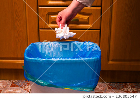 Hand placing used piece of paper into trash bin. Showcases proper waste segregation and environmental responsibility. Responsible recycling in home interior 136803547