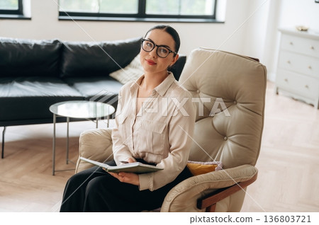 Relaxing on the chair, sitting. Woman in formal clothes is in the modern big room Relaxing on the chair, sitting. Woman in formal clothes is in the modern big room 136803721