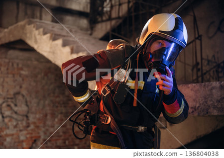 Adjusting air flow into the mask. Firefighter in professional equipment is indoors in abandoned building Adjusting air flow into the mask. Firefighter in professional equipment is indoors in abandoned building 136804038