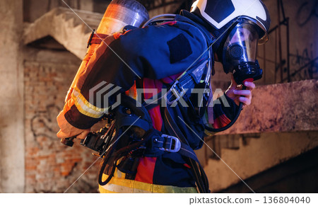 View from the side, with air balloons and mask. Firefighter in professional equipment is indoors in abandoned building View from the side, with air balloons and mask. Firefighter in professional equipment is indoors in abandoned building 136804040