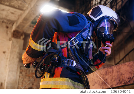 View from the side, with air balloons and mask. Firefighter in professional equipment is indoors in abandoned building View from the side, with air balloons and mask. Firefighter in professional equipment is indoors in abandoned building 136804041