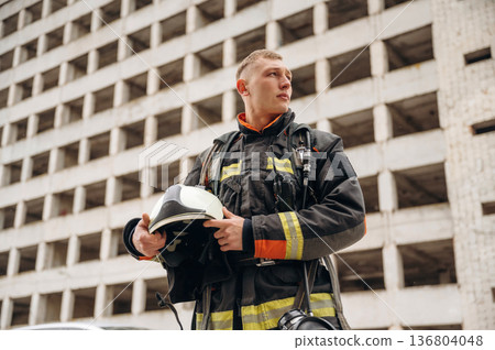 Many of the windows behind him. Professional firefighter is standing outdoors against big unfinished building 136804048