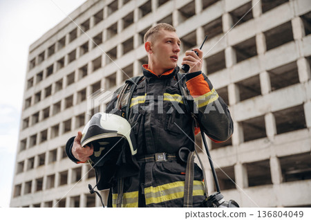 Conversation by walkie talkie. Professional firefighter is standing outdoors against big unfinished building 136804049