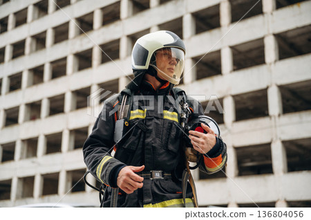 Professional firefighter is standing outdoors against big unfinished building Professional firefighter is standing outdoors against big unfinished building 136804056