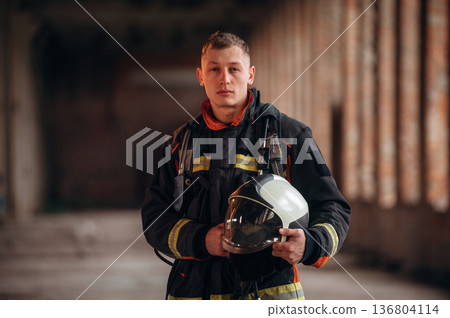 Holding helmet. Firefighter in professional equipment is indoors in abandoned building Holding helmet. Firefighter in professional equipment is indoors in abandoned building 136804114