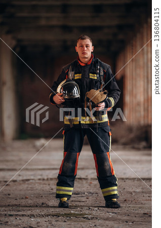 Holding helmet. Firefighter in professional equipment is indoors in abandoned building 136804115
