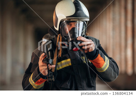 Protective air mask is on the face. Firefighter in professional equipment is indoors in abandoned building Protective air mask is on the face. Firefighter in professional equipment is indoors in abandoned building 136804134
