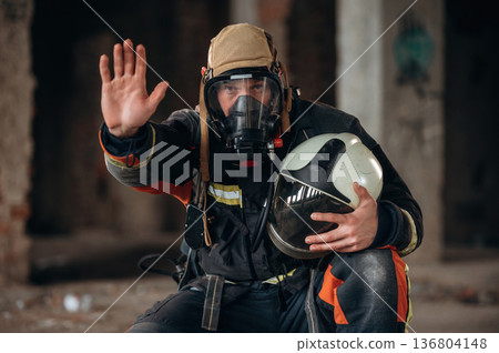 Stop hand gesture. Firefighter in professional equipment is indoors in abandoned building Stop hand gesture. Firefighter in professional equipment is indoors in abandoned building 136804148