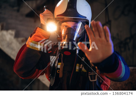 Flashlight, stop hand gesture. Firefighter in professional equipment is indoors in abandoned building 136804174