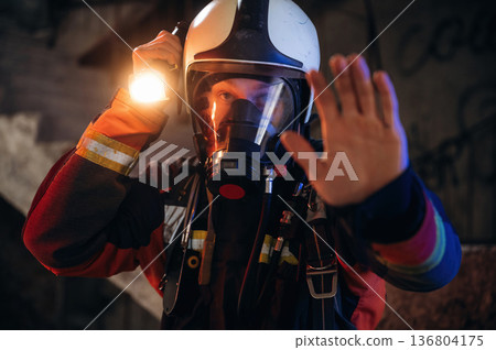 Flashlight, stop hand gesture. Firefighter in professional equipment is indoors in abandoned building 136804175