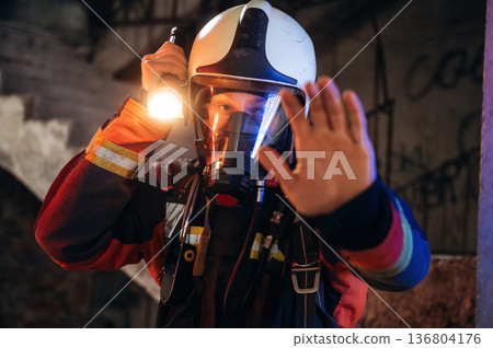Flashlight, stop hand gesture. Firefighter in professional equipment is indoors in abandoned building 136804176