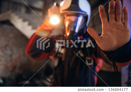 Flashlight, stop hand gesture. Firefighter in professional equipment is indoors in abandoned building Flashlight, stop hand gesture. Firefighter in professional equipment is indoors in abandoned building 136804177
