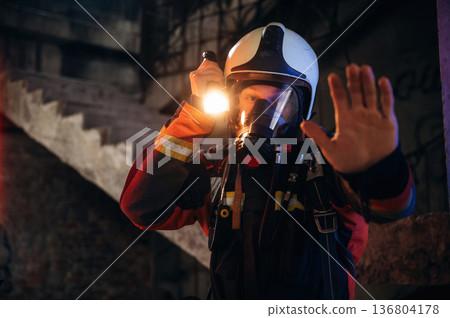 Flashlight, stop hand gesture. Firefighter in professional equipment is indoors in abandoned building Flashlight, stop hand gesture. Firefighter in professional equipment is indoors in abandoned building 136804178