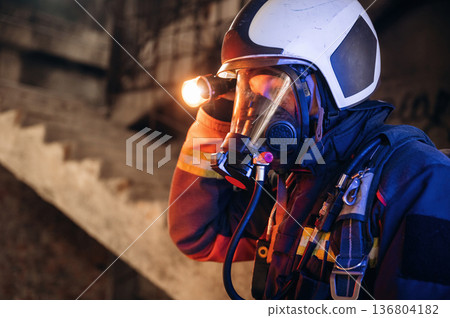 Side view, looking for the way with flashlight. Firefighter in professional equipment is indoors in abandoned building Side view, looking for the way with flashlight. Firefighter in professional equipment is indoors in abandoned building 136804182