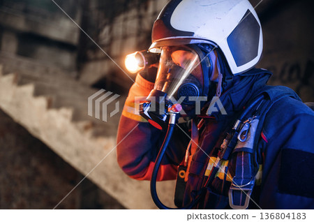 Side view, looking for the way with flashlight. Firefighter in professional equipment is indoors in abandoned building Side view, looking for the way with flashlight. Firefighter in professional equipment is indoors in abandoned building 136804183