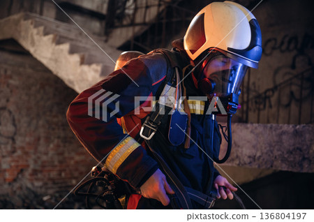 View from the side, with air balloons and mask. Firefighter in professional equipment is indoors in abandoned building View from the side, with air balloons and mask. Firefighter in professional equipment is indoors in abandoned building 136804197