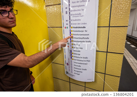 Berlin, germany, august 15,2023. Young man with glasses pointing at berlin u-bahn map on friedrichstrabe platform, checking route and planning his urban journey 136804201