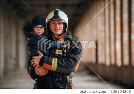 Holding kid in hands. Firefighter is with baby boy indoors Holding kid in hands. Firefighter is with baby boy indoors 136804206