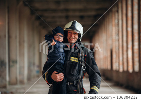 Modern hero is holding kid. Firefighter is with baby boy indoors Modern hero is holding kid. Firefighter is with baby boy indoors 136804212