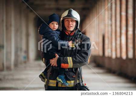 Modern hero is holding kid. Firefighter is with baby boy indoors Modern hero is holding kid. Firefighter is with baby boy indoors 136804213